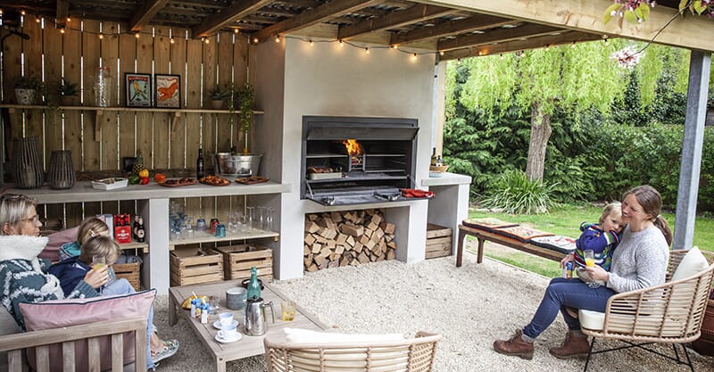 Cosy, rustic outdoor kitchen area designed by Keltie & Clark, showcasing a wood-fired oven and natural wooden furnishings in the Cotswolds.